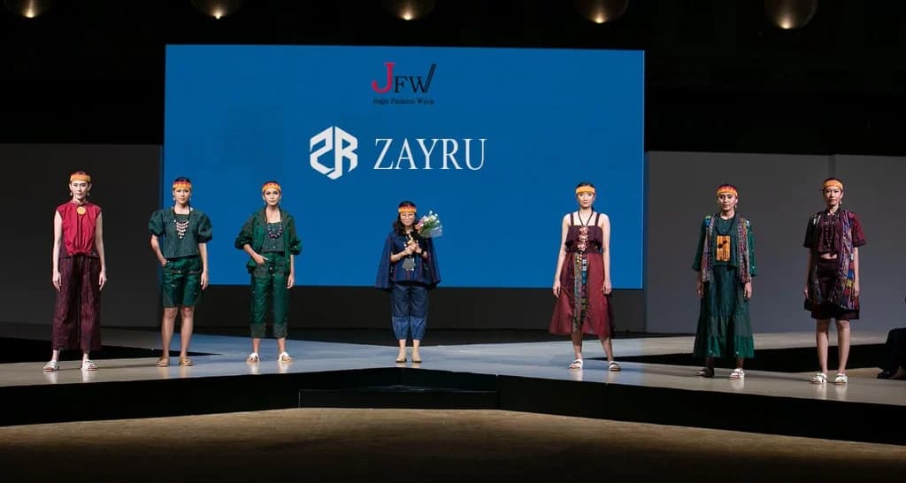 Zayru runway in front of a sky-blue LED backdrop — six models in white linen co-ords layered with indigo grid-print batik vests and skirts.